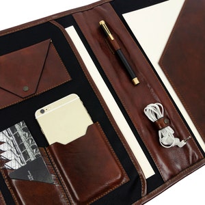 May include: A brown leather portfolio with multiple pockets and compartments. The portfolio is open and has a pen, phone, and headphones inside.