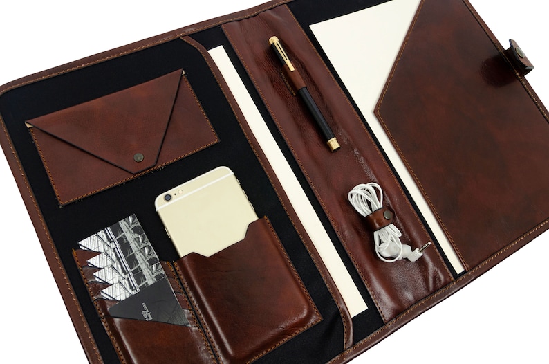 May include: A brown leather portfolio with multiple pockets and compartments. The portfolio is open and has a pen, phone, and headphones inside.