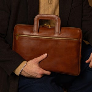 Könnte beinhalten: Eine rechteckige Aktentasche aus braunem Leder mit einem strukturierten Tragegriff, einer horizontalen Reißverschlusstasche auf der Vorderseite und goldfarbenen Metallbeschlägen. Die Tasche hat eine glatte Oberfläche und sichtbare Nähte.