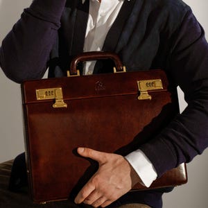 May include: A dark brown leather briefcase featuring a structured rectangular shape, a sturdy top handle, and two gold-toned combination locks on the front. The surface has a smooth, polished finish with visible stitching along the edges.