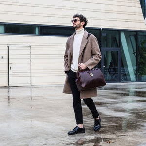 May include: A man wearing a brown coat, black pants, and black shoes is walking outside. He is carrying a brown leather briefcase with a gold lock.