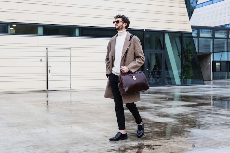 May include: A man wearing a brown coat, black pants, and black shoes is walking outside. He is carrying a brown leather briefcase with a gold lock.