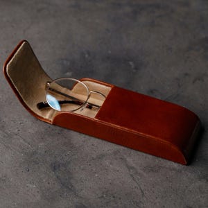 May include: A smooth, cognac-colored leather glasses case with a curved, open flap revealing a soft, beige interior lining. A pair of round-framed eyeglasses with thin metal temples rests partially inside the open case, showing clear lenses.