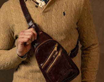 Puede incluir: Un bolso bandolera de cuero marrón oscuro con una correa ajustable, dos compartimentos frontales con cremallera y herrajes dorados, y un pequeño logotipo grabado en la parte inferior, llevado cruzado sobre el pecho con un suéter de punto.