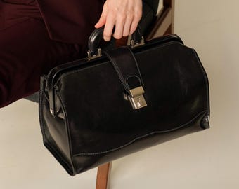 May include: A black leather doctor's bag with a silver lock and handle. The bag is sitting on a wooden chair.
