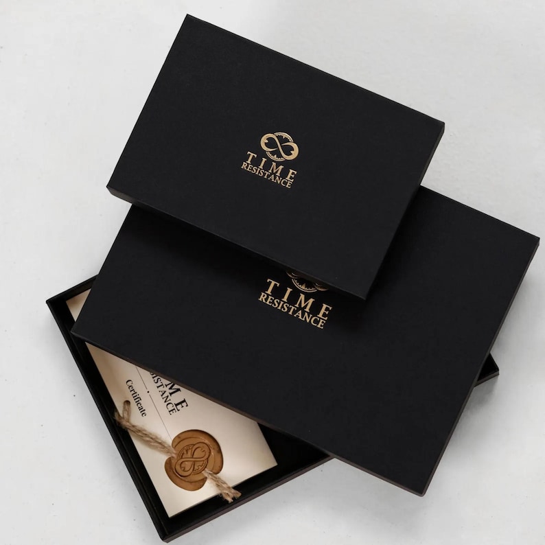 May include: Two rectangular black gift boxes stacked, featuring the gold logo and text TIME RESISTANCE. The bottom box is partially open, revealing a cream-colored certificate with a brown wax seal attached by a piece of twine.