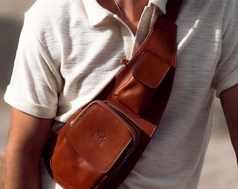 May include: A tan leather sling bag featuring a main zippered compartment with a flap pocket on the front and a smaller vertical pocket on the shoulder strap. The bag displays a small embossed logo on the front pocket and has visible stitching.