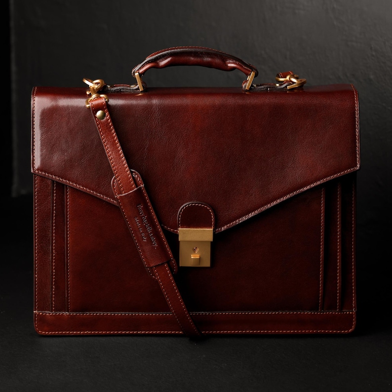 May include: A brown leather briefcase with a gold clasp and a shoulder strap. The strap has the text "Genuine Leather" embossed on it.