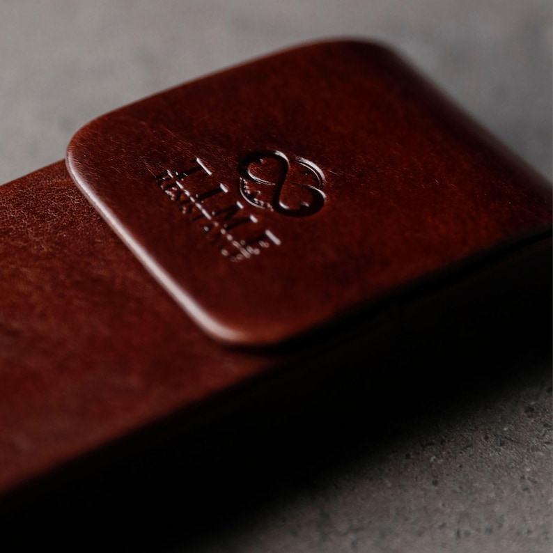 May include: A dark brown leather case with a smooth, rounded rectangular flap. The surface features a debossed logo consisting of an infinity symbol above the text "TIME" in a clean, sans-serif font, centered on the flap's top section.