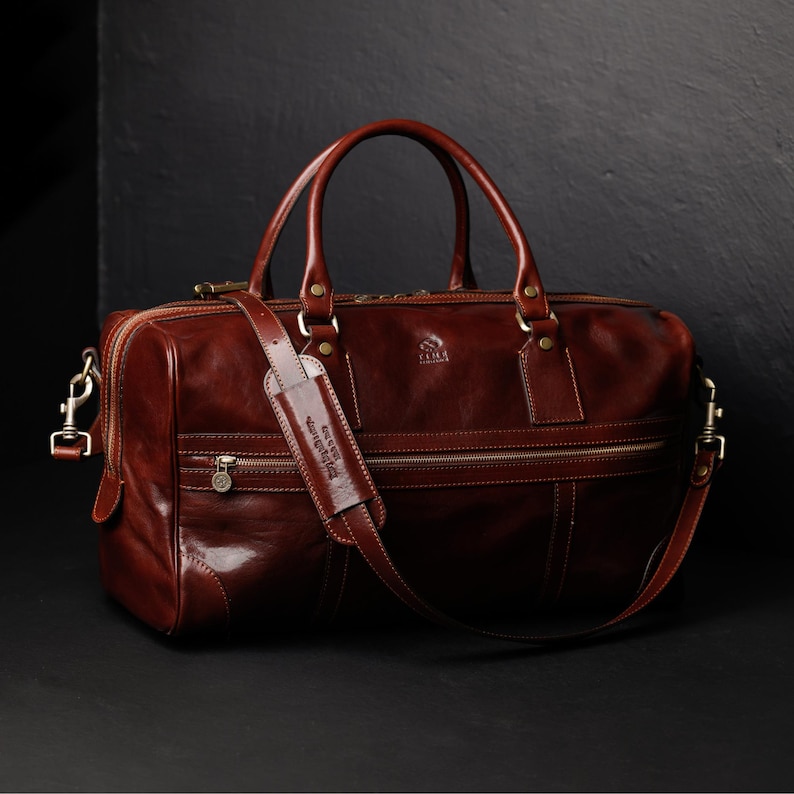 K&ouml;nnte beinhalten: Eine braune Reisetasche aus Leder mit einem oberen Griff und einem Schultergurt. Die Tasche hat einen Rei&szlig;verschluss und eine kleine Tasche auf der Vorderseite. Die Tasche hat ein Markenetikett mit der Aufschrift "ILMA" auf der Vorderseite.