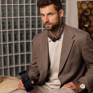 May include: A tan blazer worn over a cream-colored ribbed sweater and a black patterned neckerchief. A black leather case with a small embossed logo is held in one hand, and a wristwatch with a white face and brown leather strap is visible.