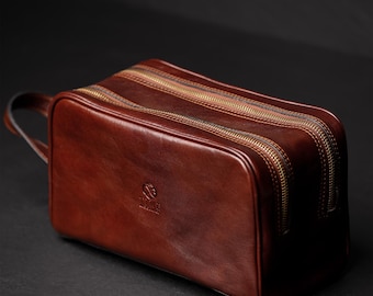 May include: A rectangular brown leather toiletry bag featuring two separate zippered compartments with metallic gold-toned zippers. A small leather handle is attached to the side, and a small embossed logo is visible on the front surface.