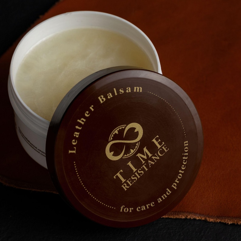 May include: A white plastic jar containing a pale yellow leather balsam. The dark brown lid features the gold text Leather Balsam, TIME RESISTANCE, and for care and protection, along with an infinity symbol logo centered on the circular surface.