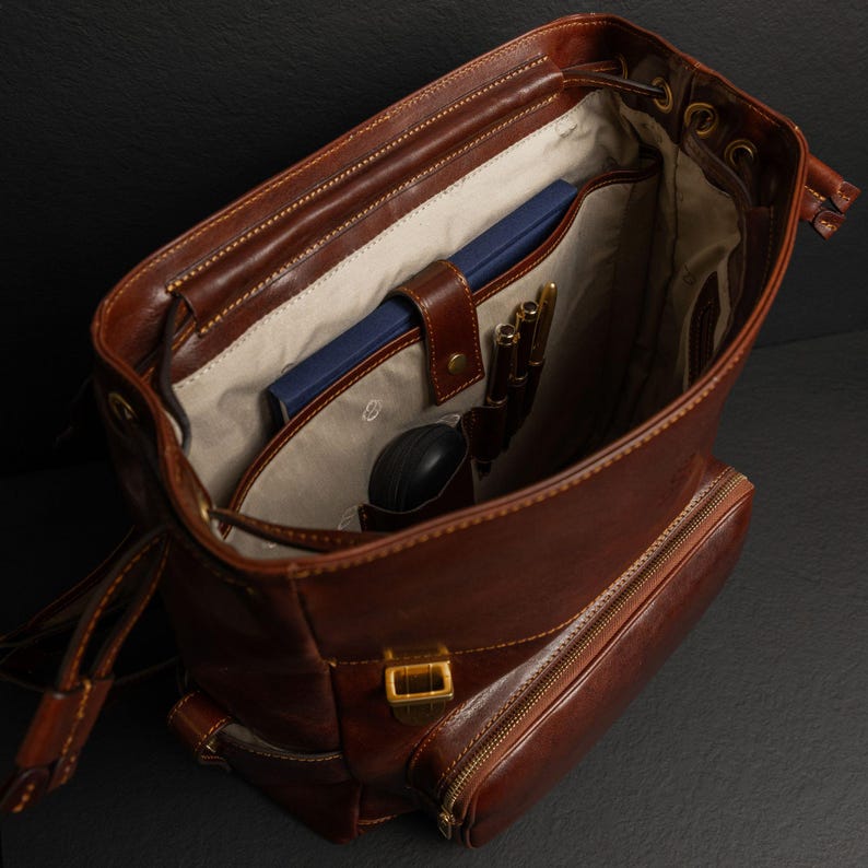 K&ouml;nnte beinhalten: Ein brauner Lederrucksack mit Kordelzugverschluss und einer Rei&szlig;verschlusstasche vorne. Das Innere ist mit hellbeigem Stoff ausgekleidet und verf&uuml;gt &uuml;ber ein Fach mit einem blauen Notizbuch, einem kleinen schwarzen Etui und zwei goldenen Stiften.