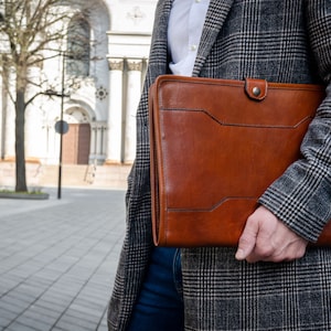 May include: A brown leather portfolio with a flap closure and a metal button. The portfolio is held by a person wearing a gray plaid coat.