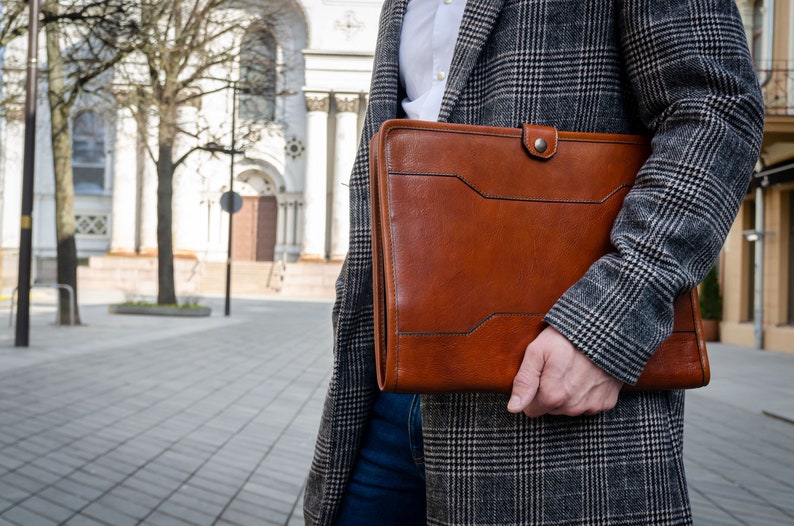 May include: A brown leather portfolio with a flap closure and a metal button. The portfolio is held by a person wearing a gray plaid coat.