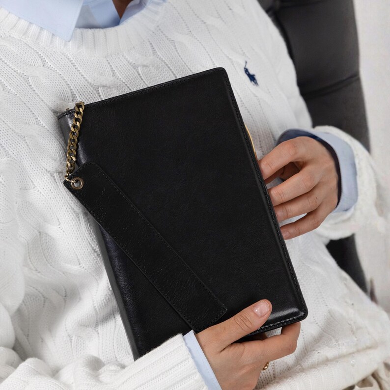 May include: A rectangular black leather clutch featuring a smooth finish and a gold-toned metal chain attachment connected to a matching black leather wrist strap. The clutch has a structured design with clean edges and a minimalist aesthetic.