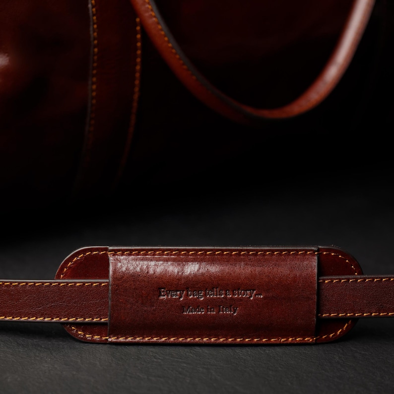 K&ouml;nnte beinhalten: Nahaufnahme eines braunen Lederriemens f&uuml;r eine Tasche mit dem gepr&auml;gten Text "Every bag tells a story... Made in Italy" auf dem Riemen.