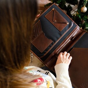 May include: A brown leather portfolio with a black canvas interior and multiple pockets. The portfolio is open and the person is holding it with their right hand.