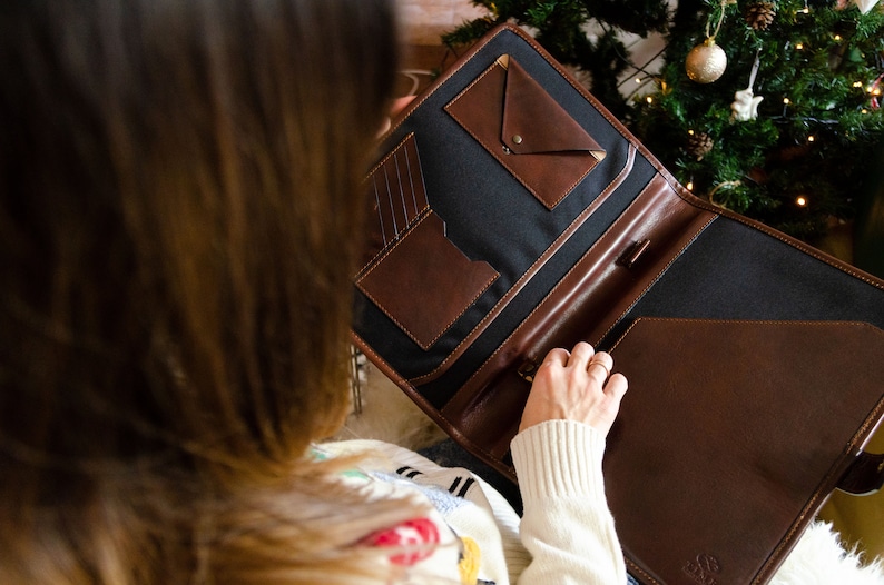 May include: A brown leather portfolio with a black canvas interior and multiple pockets. The portfolio is open and the person is holding it with their right hand.