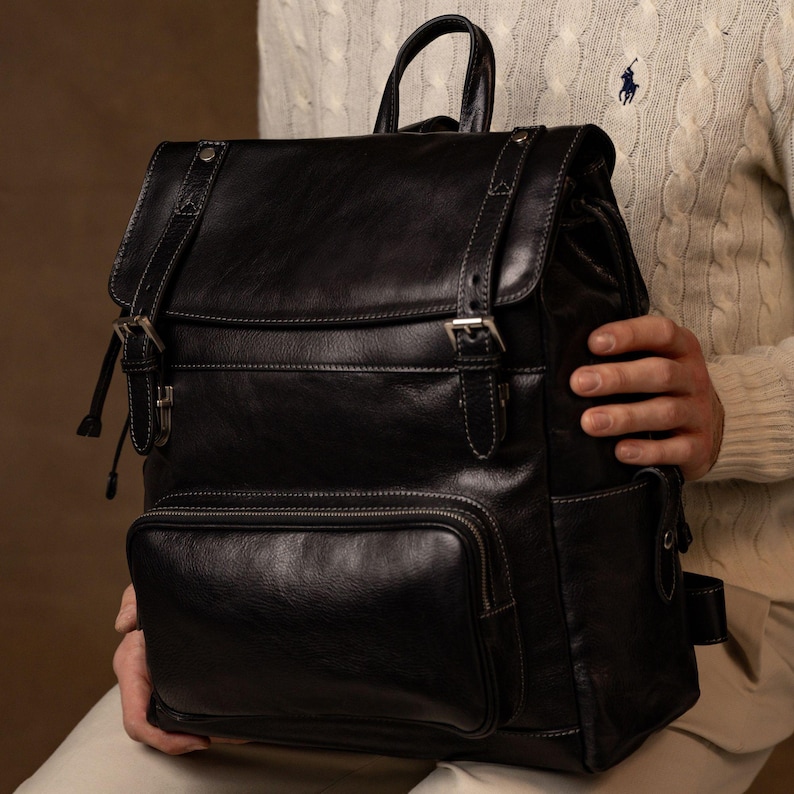 K&ouml;nnte beinhalten: Ein schwarzer Lederrucksack mit einer oberen Klappe mit zwei Schnallenriemen, einer vorderen Rei&szlig;verschlusstasche und einem Tragegriff. Die Tasche verf&uuml;gt &uuml;ber silberfarbene Metallbeschl&auml;ge, sichtbare N&auml;hte und Seitentaschen.
