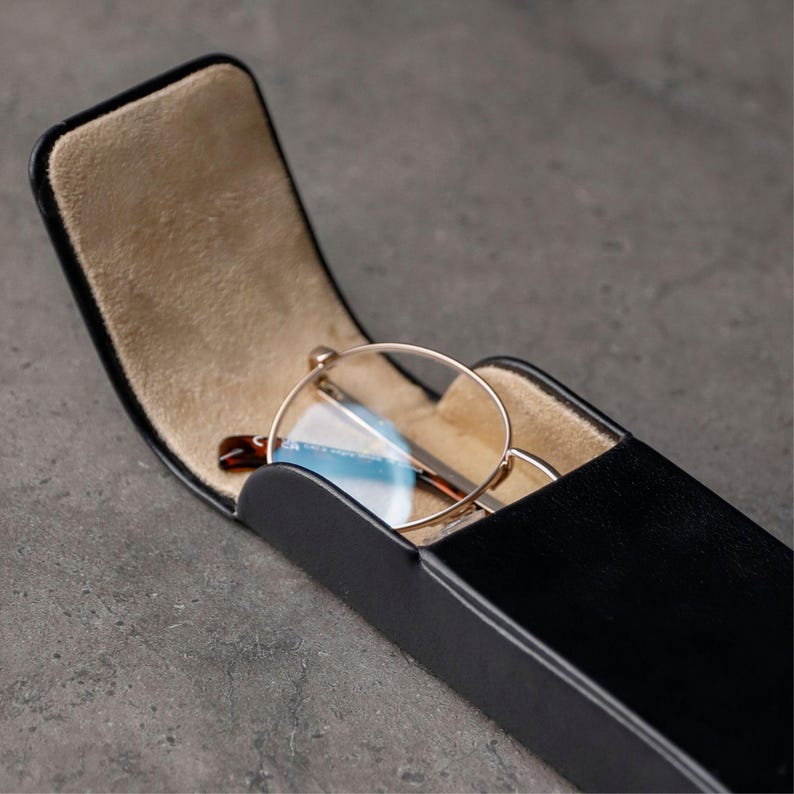 May include: A black rectangular glasses case with a hinged lid, featuring a smooth exterior and a soft beige interior lining. Inside the open case rests a pair of round-framed eyeglasses with thin gold-tone metal rims and clear lenses.