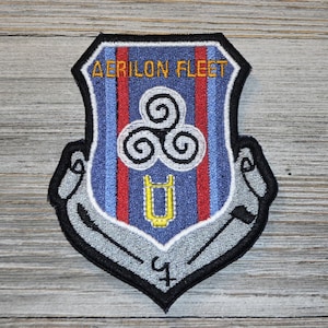 May include: A gray and black embroidered patch with a blue, red, and white shield. The shield has a white symbol with three spirals and a yellow symbol that looks like a tuning fork. The text "AERILON FLEET" is above the shield in yellow.