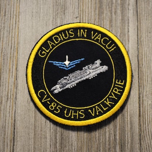 May include: A black and yellow embroidered patch with the text "GLADIUS IN VACUI CV-85 UHS VALKYRIE" and a white silhouette of a spaceship with a blue and white wing design.