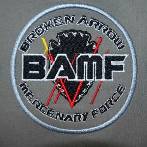 Può includere: Toppa ricamata circolare con la scritta "BROKEN ARROW MERCENARY FORCE". La toppa presenta una punta di freccia nera, le lettere "BAMF" e dettagli rossi e gialli su sfondo grigio.