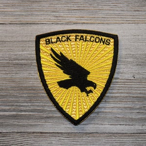 以下が含まれることがあります： 黄色い背景に黒いファルコンのシルエットが刺繍された黒いパッチで、黒い文字で「Black Falcons」と書かれています。