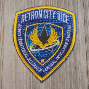 May include: A blue and yellow embroidered patch with the text "Detron City Vice" and "Aurani Territorial Alliance Central Intelligence Bureau".