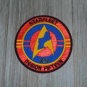 May include: A red, yellow, and blue embroidered patch with a black outline. The patch features a stylized starfleet insignia with the text "Starfleet Region Fifteen" and "XV" in the center.