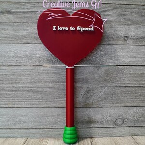 Peut inclure: Un panneau rouge en forme de c&oelig;ur avec le texte "I love to Spend" en blanc. Le panneau est fix&eacute; &agrave; un poteau rouge avec une base verte. Le fond est une surface en bois.