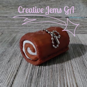Op de afbeelding: Een kleine, bruine cake in Zwitserse rolstijl met een witte, gedraaide vulling, bevestigd aan een zilveren ketting. De cake staat op een houten oppervlak. De tekst "Creative Jems GA" staat boven de cake.