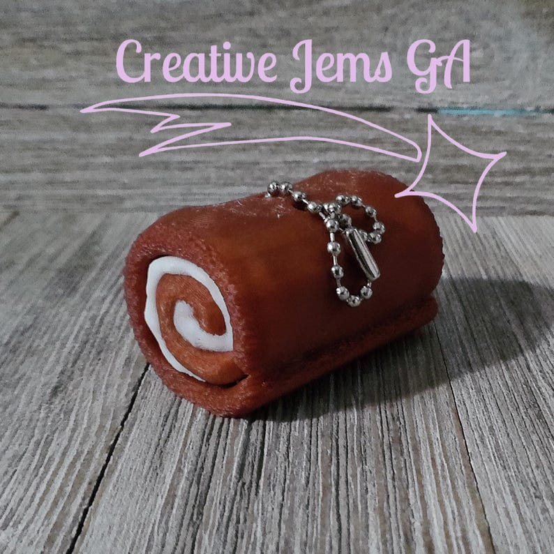 Op de afbeelding: Een kleine, bruine cake in Zwitserse rolstijl met een witte, gedraaide vulling, bevestigd aan een zilveren ketting. De cake staat op een houten oppervlak. De tekst "Creative Jems GA" staat boven de cake.
