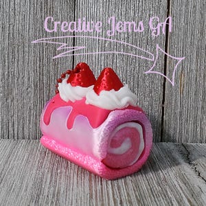 Op de afbeelding: Een roze en witte decoratieve cake roll met aardbeientoppings en slagroom. De cake roll heeft een wervelontwerp en is versierd met rode aardbeivormige accenten. De afbeelding bevat de tekst "Creative Jems Gift".