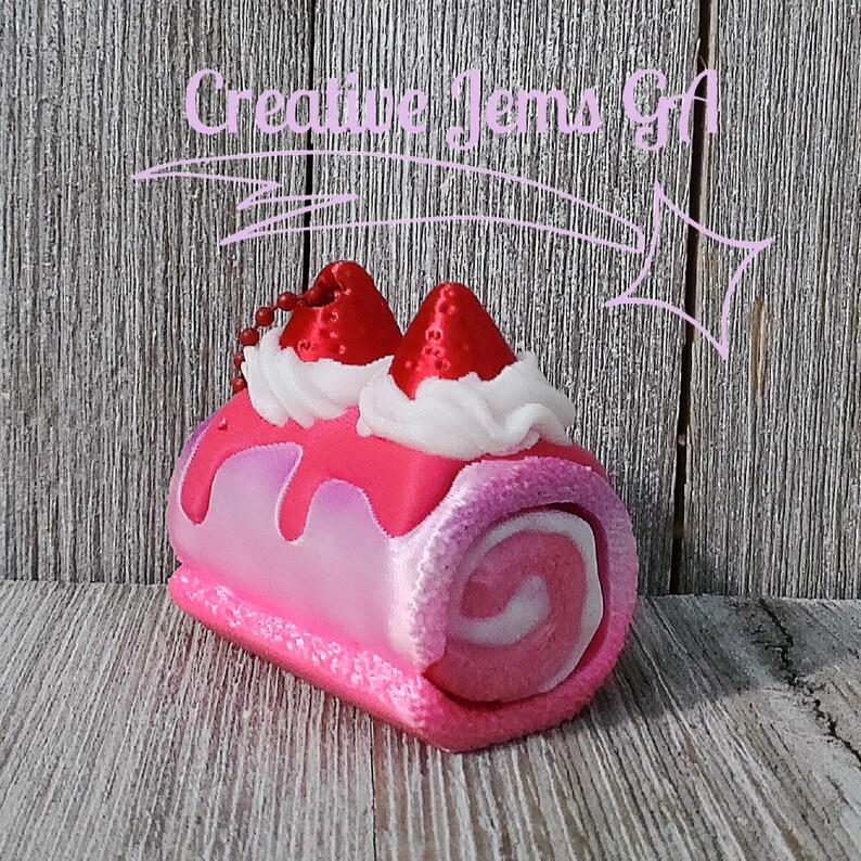 Op de afbeelding: Een roze en witte decoratieve cake roll met aardbeientoppings en slagroom. De cake roll heeft een wervelontwerp en is versierd met rode aardbeivormige accenten. De afbeelding bevat de tekst "Creative Jems Gift".