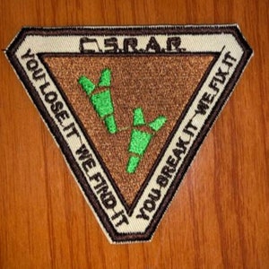 May include: A triangular patch with a brown background and a white border. The patch has green stylized footprints and the text "You lose it we find it - You break it we fix it" around the border. The text "S.R.A.F." is in the top centre of the patch.