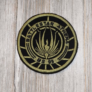 May include: A round, black and gold embroidered patch with the text "Battlestar Athena" and "BS 95" in a circular design. The patch features a stylised, black and gold, winged symbol.