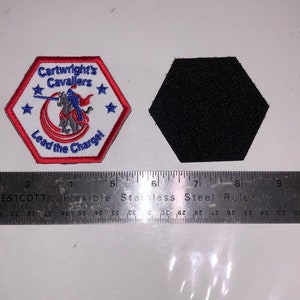 May include: A white hexagonal patch with red and blue embroidered details. The patch features a knight riding a horse, a crescent moon, and stars. The text "Cartwright's Cavaliers" and "Lead the Charge!" is embroidered on the patch.
