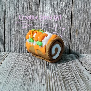 Op de afbeelding: Een decoratieve, vilten Zwitserse rolcake met oranje glazuur, witte vulling en groene en gele versieringen. De cake staat tegen een rustieke houten achtergrond. De tekst "Creative Items Gift" staat boven de cake.