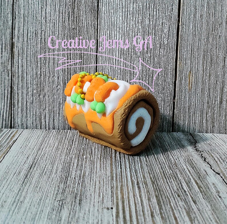 Op de afbeelding: Een decoratieve, vilten Zwitserse rolcake met oranje glazuur, witte vulling en groene en gele versieringen. De cake staat tegen een rustieke houten achtergrond. De tekst "Creative Items Gift" staat boven de cake.