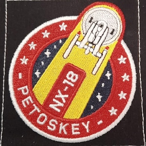 May include: A red and white embroidered patch with a yellow spaceship and the text "NX-18 Petoskey" in white.