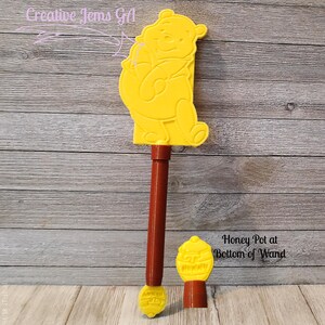Peut inclure: Une baguette jaune en forme de Winnie l'ourson avec un manche marron et un pot de miel en bas. La baguette porte le texte "Honey Pot at Bottom of Wand". La baguette est en plastique.