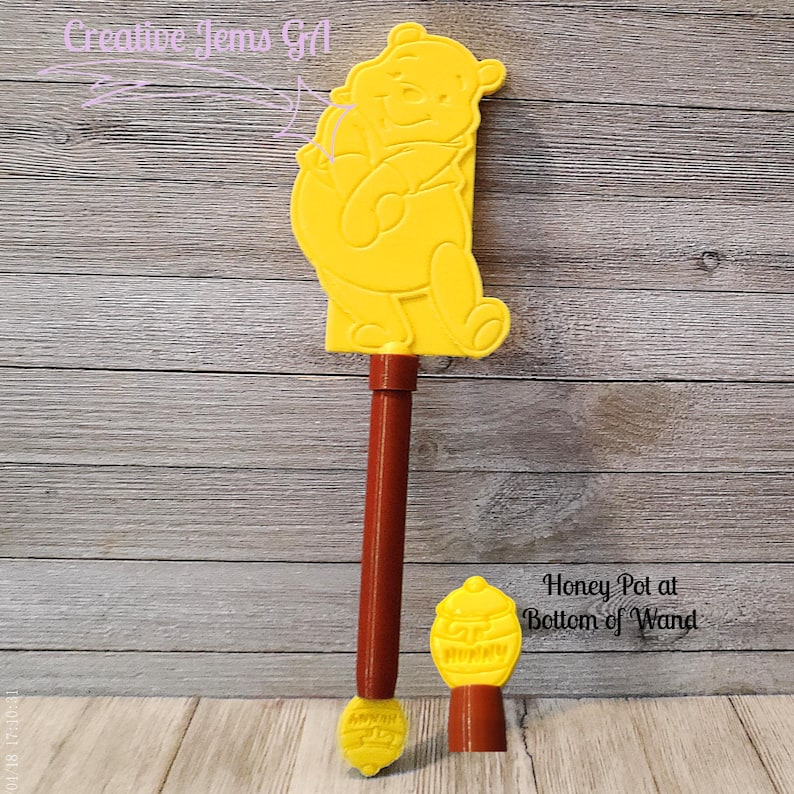 Peut inclure: Une baguette jaune en forme de Winnie l'ourson avec un manche marron et un pot de miel en bas. La baguette porte le texte "Honey Pot at Bottom of Wand". La baguette est en plastique.