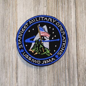 May include: A blue and black embroidered patch with the text "Starfleet Military Operations USS Iwo Jima" around the edge. The center of the patch features a white mountain with a flag planted on top, and three green figures climbing the mountain.