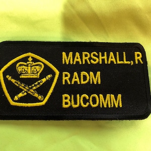 May include: Black embroidered patch with a gold crown and crossed pipes. The text "MARSHALL, R RADM BUCOMM" is embroidered in gold.