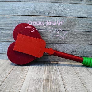 Peut inclure: Un objet rouge et vert en forme de c&oelig;ur avec un dessus rectangulaire et une longue poign&eacute;e. La poign&eacute;e est rouge avec une poign&eacute;e verte. L'objet est sur une surface en bois. Le texte "Creative Gems GA" est en arri&egrave;re-plan.