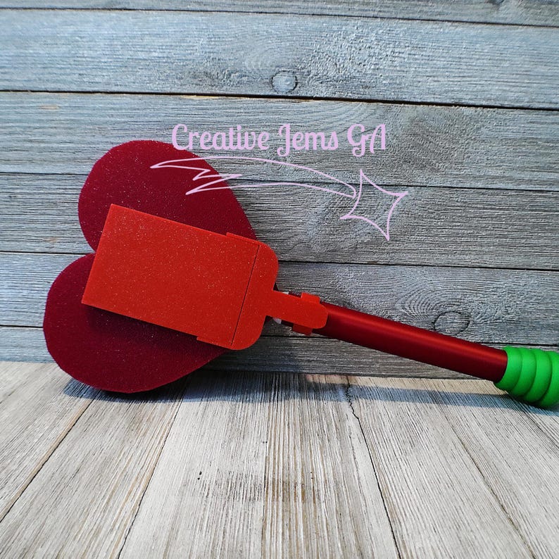 Peut inclure: Un objet rouge et vert en forme de c&oelig;ur avec un dessus rectangulaire et une longue poign&eacute;e. La poign&eacute;e est rouge avec une poign&eacute;e verte. L'objet est sur une surface en bois. Le texte "Creative Gems GA" est en arri&egrave;re-plan.