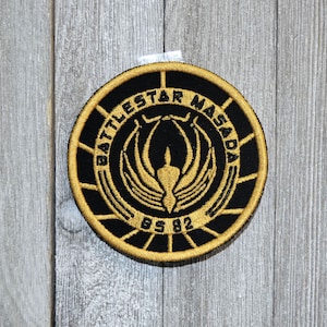 May include: A black and gold embroidered patch with the text "Battlestar Masada" and the year "BS 82" in a circle.